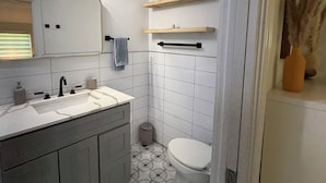 Ein Badezimmer mit einem weißen Waschbecken, Spiegel und Toilette. Über der Toilette befinden sich Holzregale und daneben ein Mülleimer.