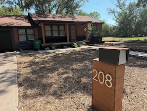 A wooden mailbox with the number 208, a building with a brown roof, and a mulched area with trees in the background. - Bright & Peaceful 3-bedroom house with AC in Westworth Village (Westworth Village)