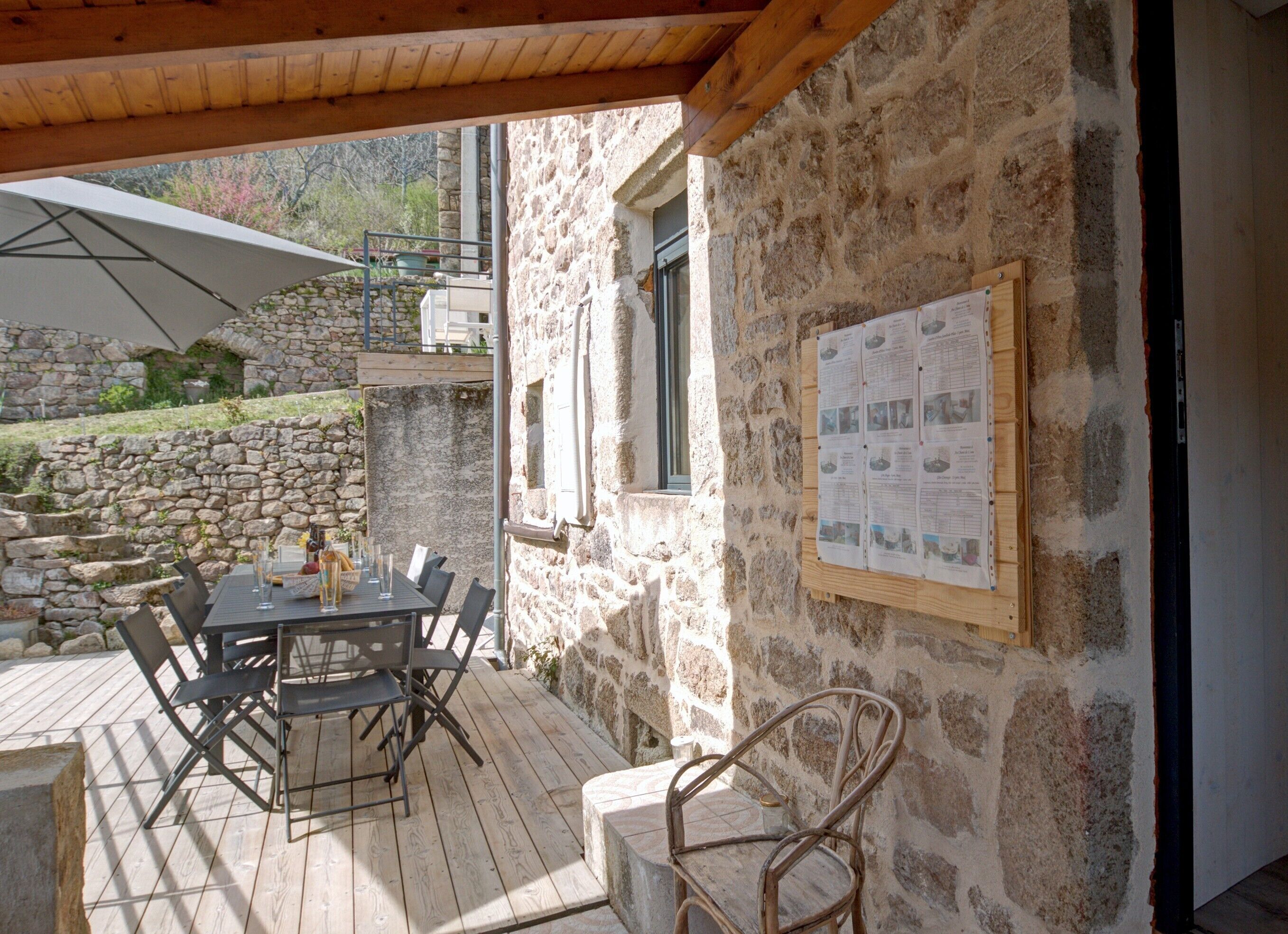 Une terrasse extérieure avec un mur en pierre, une table en bois avec des chaises et un panneau d’affichage où sont accrochés des papiers.