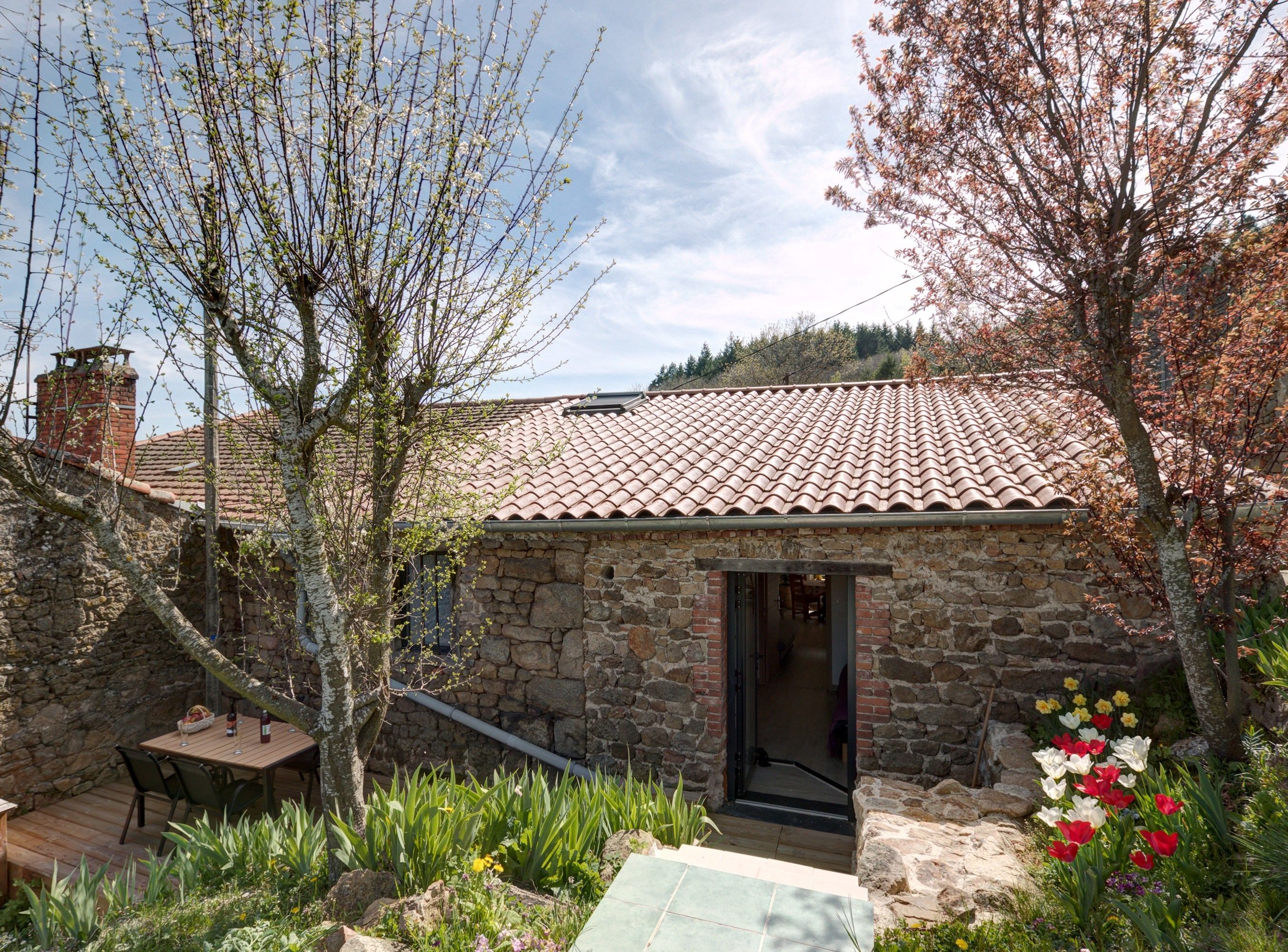 Un bâtiment en pierre avec un toit en tuiles, une terrasse en bois et un jardin fleuri.