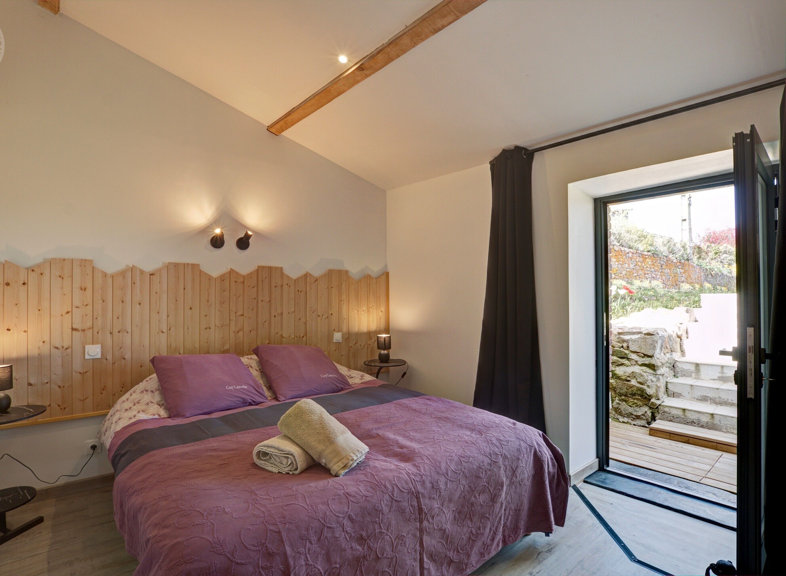 Ein Schlafzimmer mit einem großen Bett, einem Holzkopfstück, zwei Nachttischlampen und Blick durch eine Glastür auf einen Außenbereich.