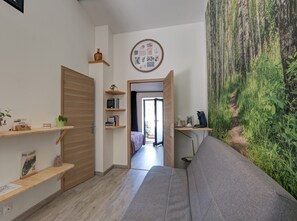 A room with a grey sofa, wooden shelves with books and a small plant, a door leading to another room, and a wall mural of a forest.