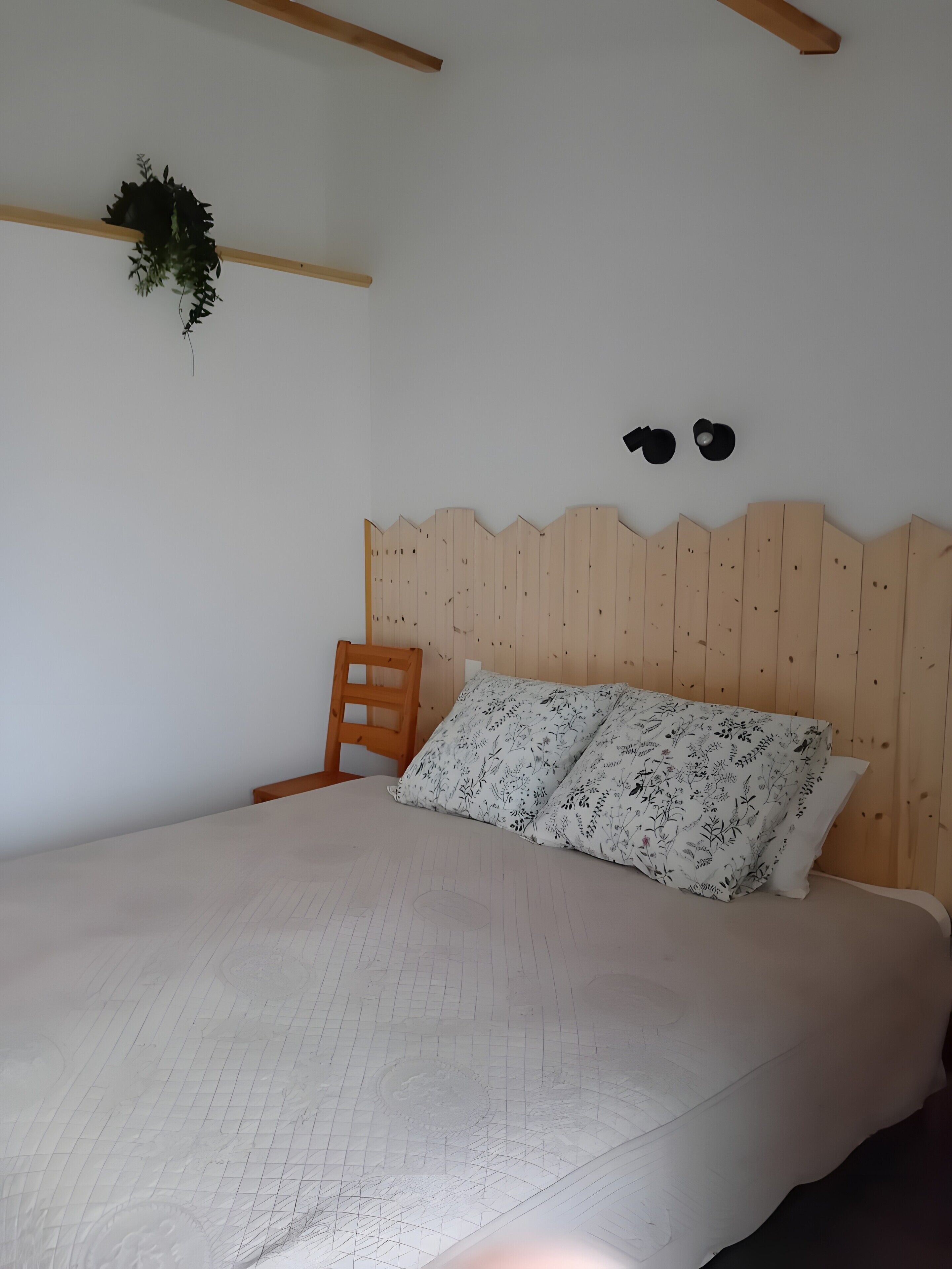 Une chambre avec une tête de lit en bois, un lit simple avec une literie à motifs floraux, une chaise en bois et une plante fixée au mur.