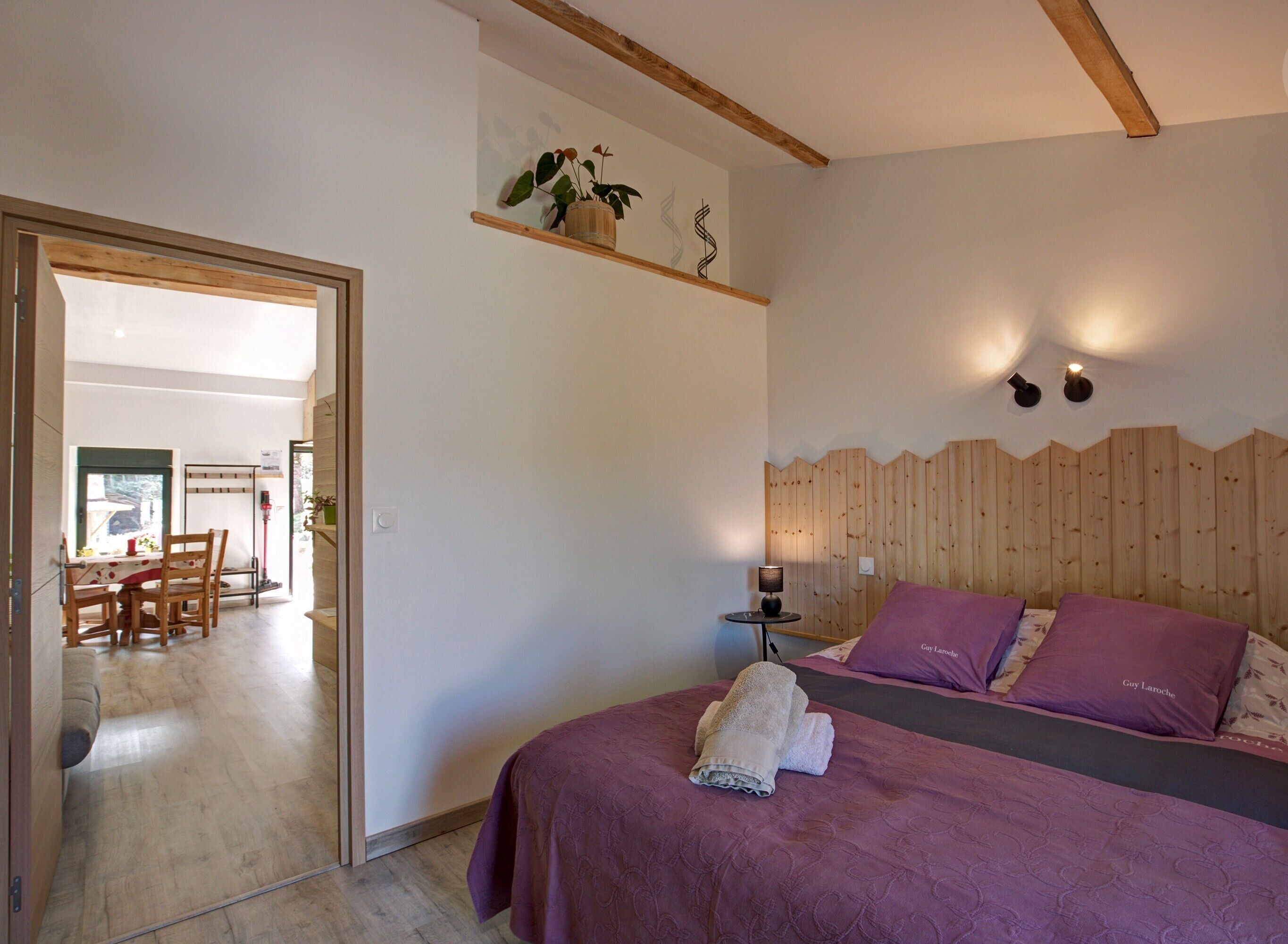 Une chambre à coucher avec une tête de lit en bois, un lit avec une literie violette, une table de chevet avec une lampe et une petite plante en pot posée sur une étagère.