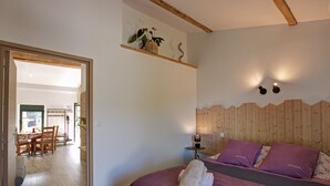 Quarto com cabeceira de madeira, cama com roupa de cama roxa, mesa de cabeceira com abajur e uma pequena planta em vaso sobre uma prateleira.