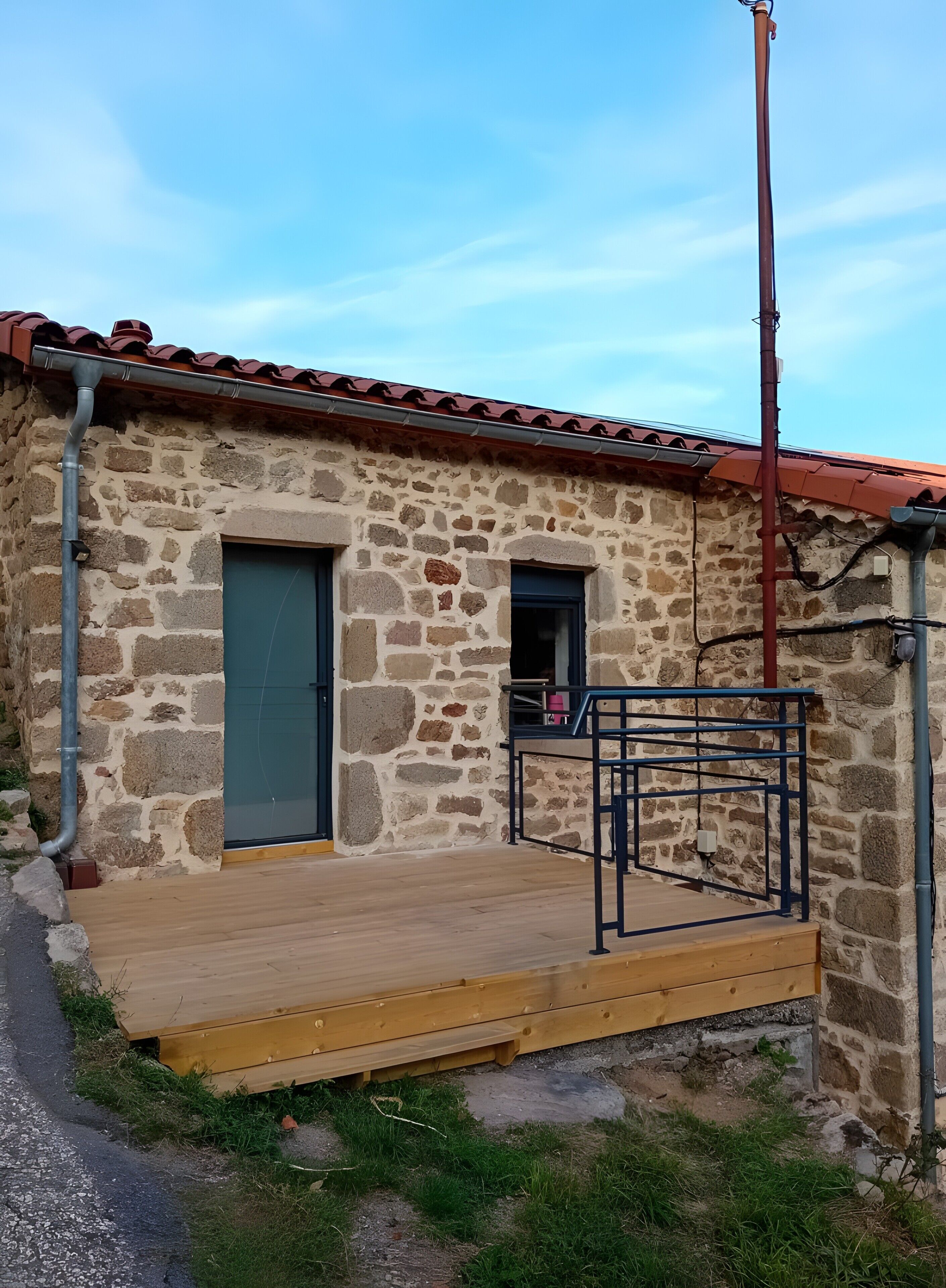 Un bâtiment en pierre doté d’une terrasse en bois, d’une rambarde en métal et d’une porte.