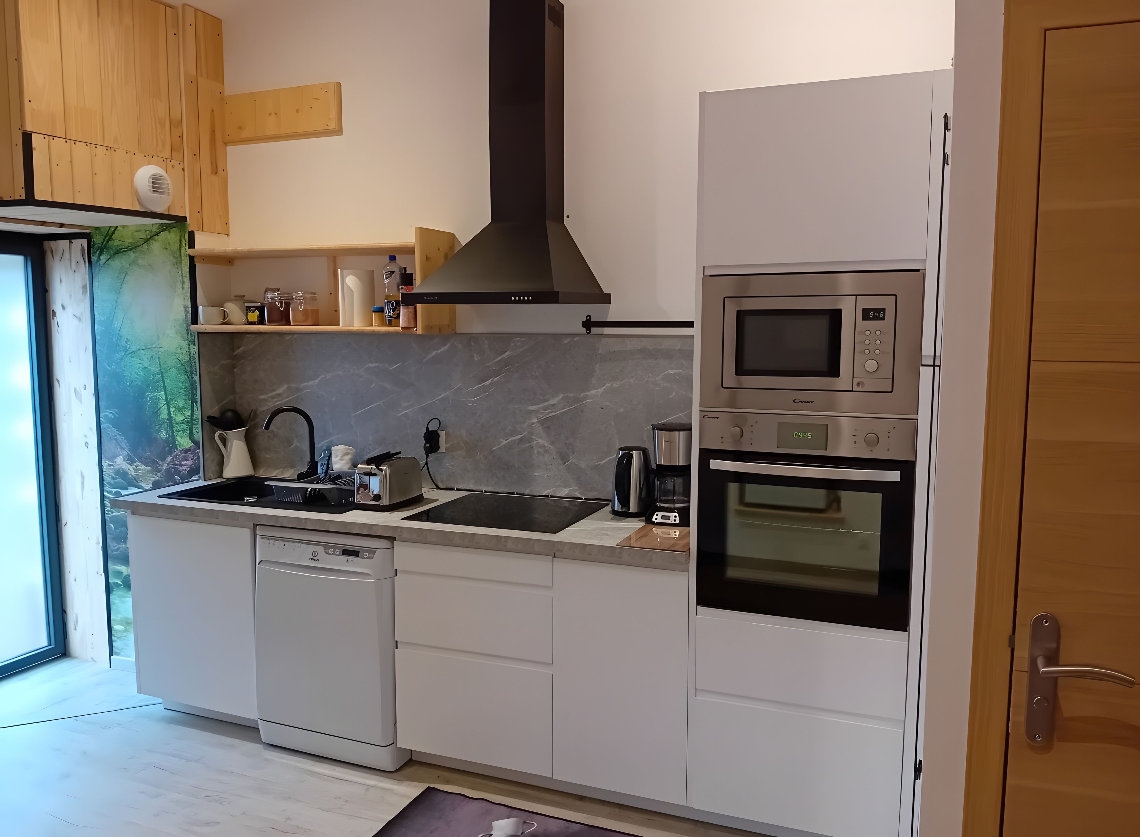 Une cuisine moderne équipée d’un lave-vaisselle, d’un four à micro-ondes et d’un four traditionnel, avec des meubles en bois et une vue sur une forêt par la fenêtre.