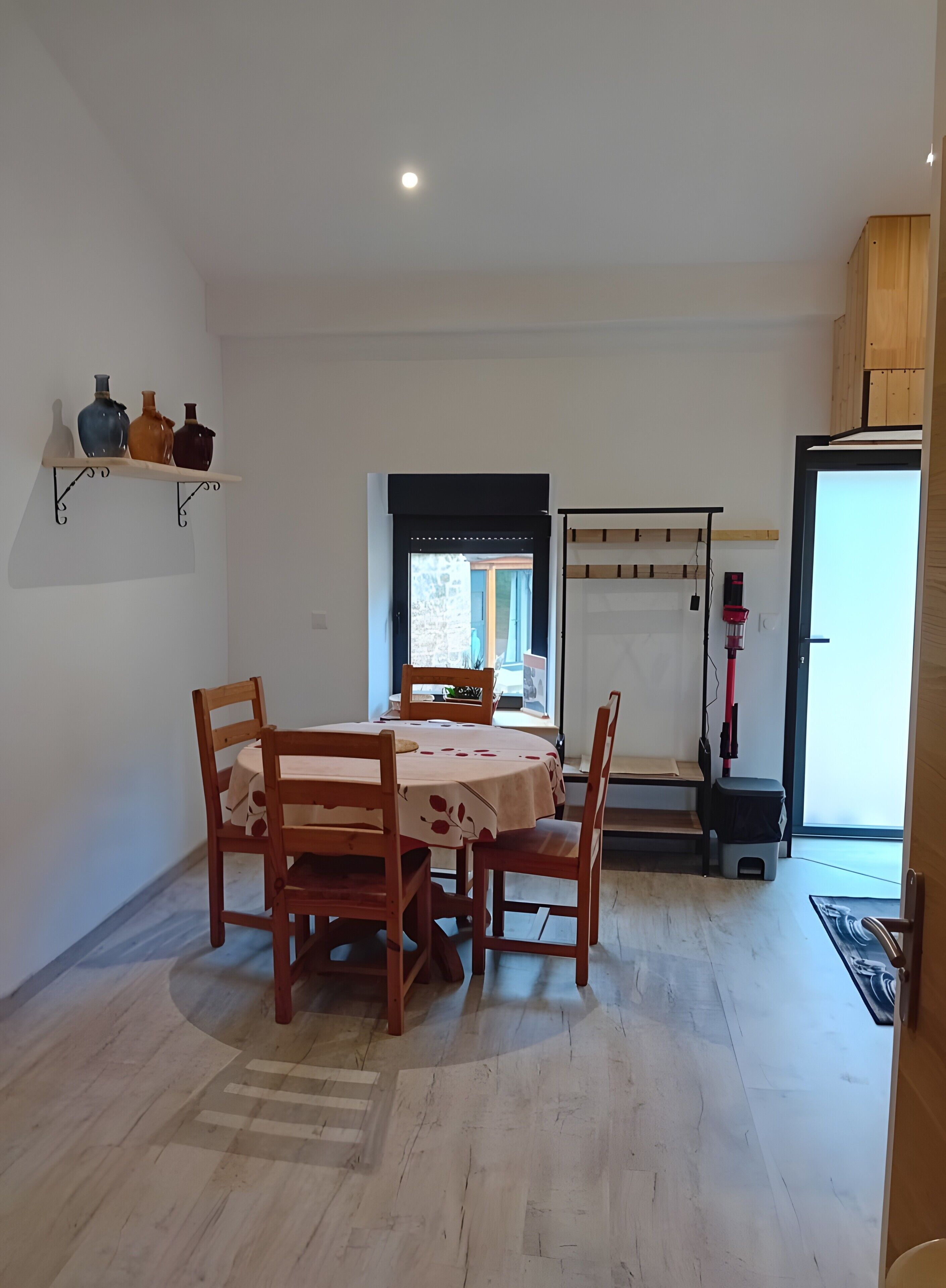 A dining area with a table and chairs, a shelf with decorative items, a window with a view, and a vacuum cleaner.