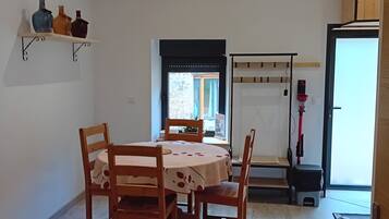 A dining area with a table and chairs, a shelf with decorative items, a window with a view, and a vacuum cleaner.