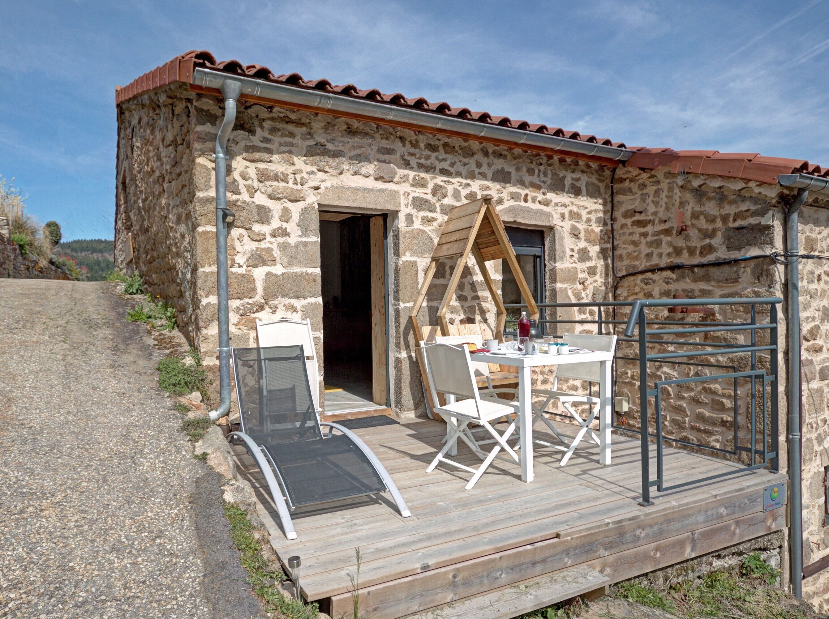 Un bâtiment en pierre doté d’une terrasse en bois, d’une table avec des chaises et d’un portail en métal.