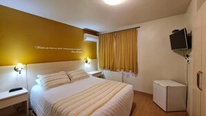 Quarto de hotel com cama, mesas de cabeceira, televisão e um pequeno frigorífico.
