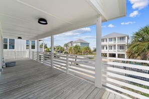 Una terraza de madera cubierta con barandas blancas, vista despejada a una propiedad frente al mar y una casa de dos pisos con balcones.