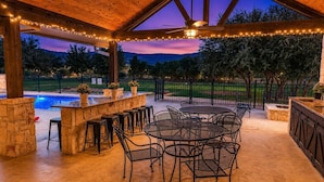 Un patio al aire libre con piscina, muros de piedra, mobiliario metálico y luces colgantes.