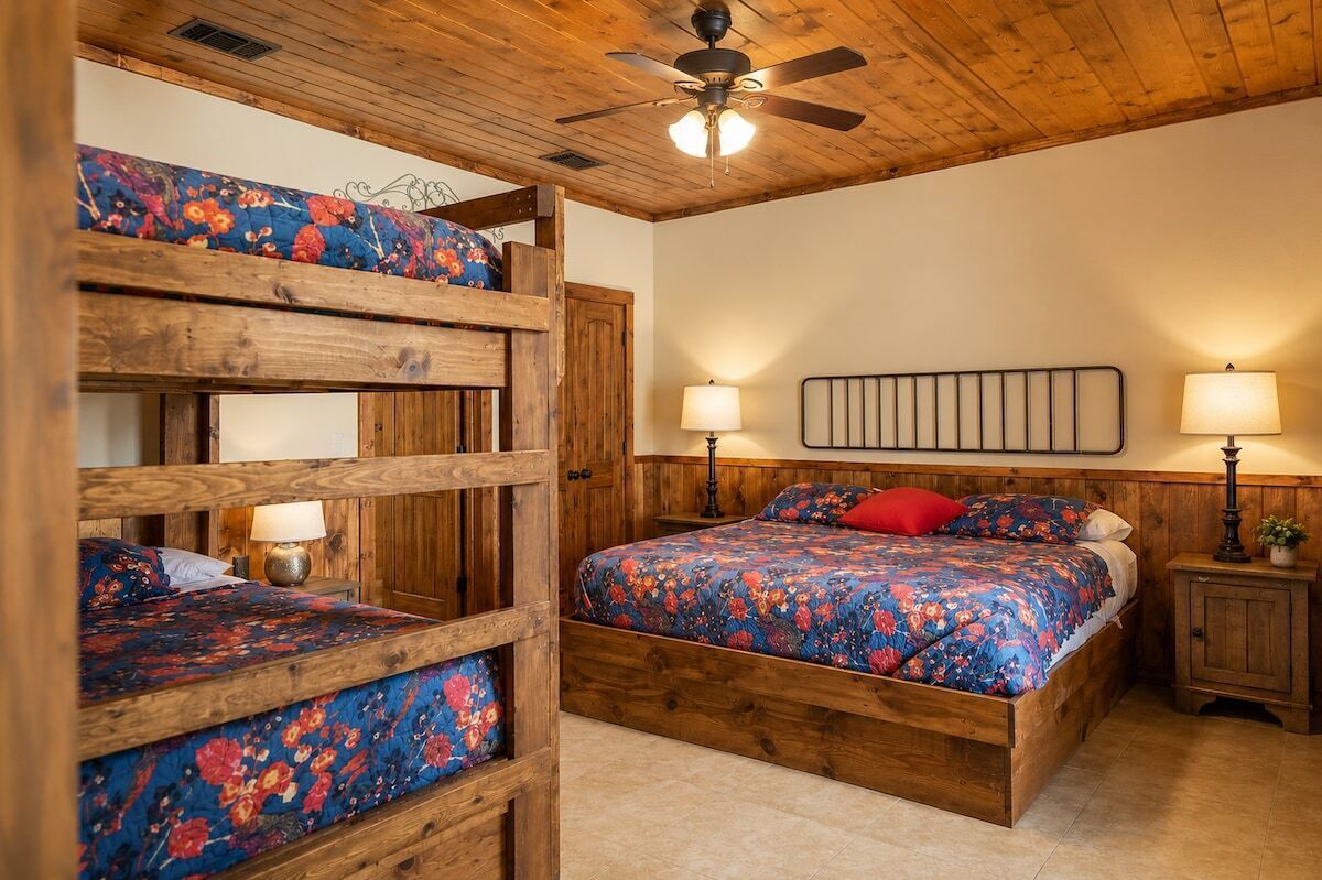 A bedroom with a bunk bed, a single bed, wooden headboard, two bedside lamps, and a ceiling fan.
