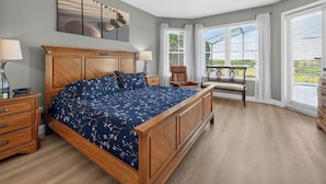 Ein Schlafzimmer mit einem großen Bett, einem Holzkopfende, einem Nachttisch, einem Sessel und Blick ins Freie durch große Fenster.