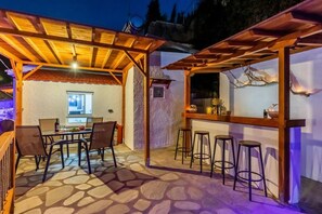 Une terrasse extérieure dotée d’un sol en pierre, d’une pergola en bois et d’un espace bar avec des tabourets.