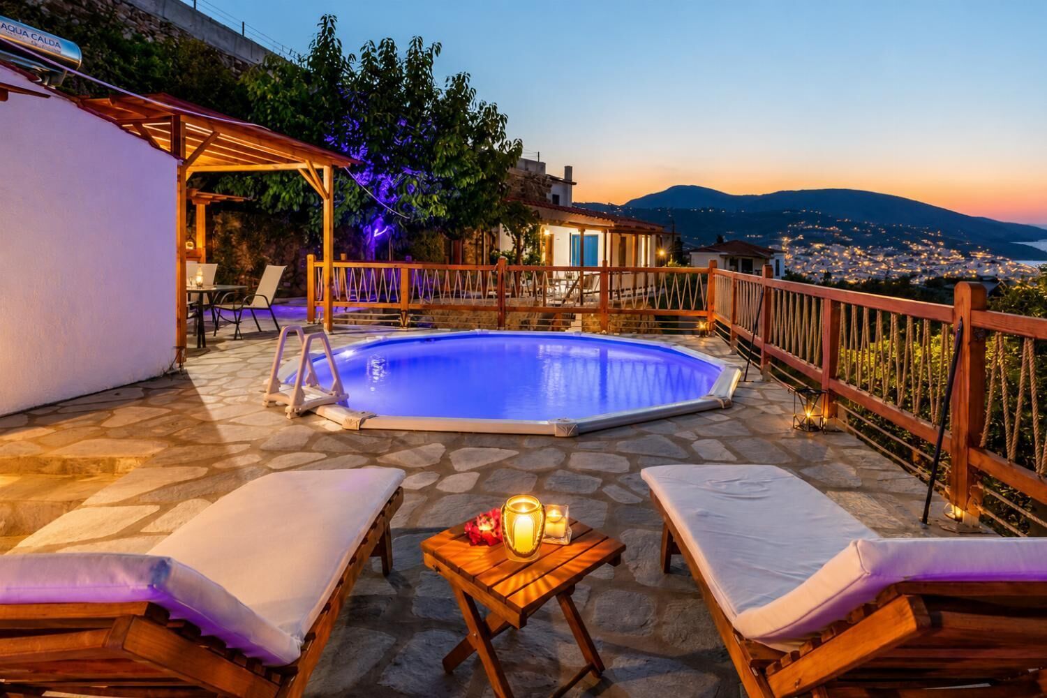 Ein Pool mit Liegestühlen und einem Tisch, umgeben von einer Holzterrasse und Blick auf eine Stadtlandschaft in der Abenddämmerung.