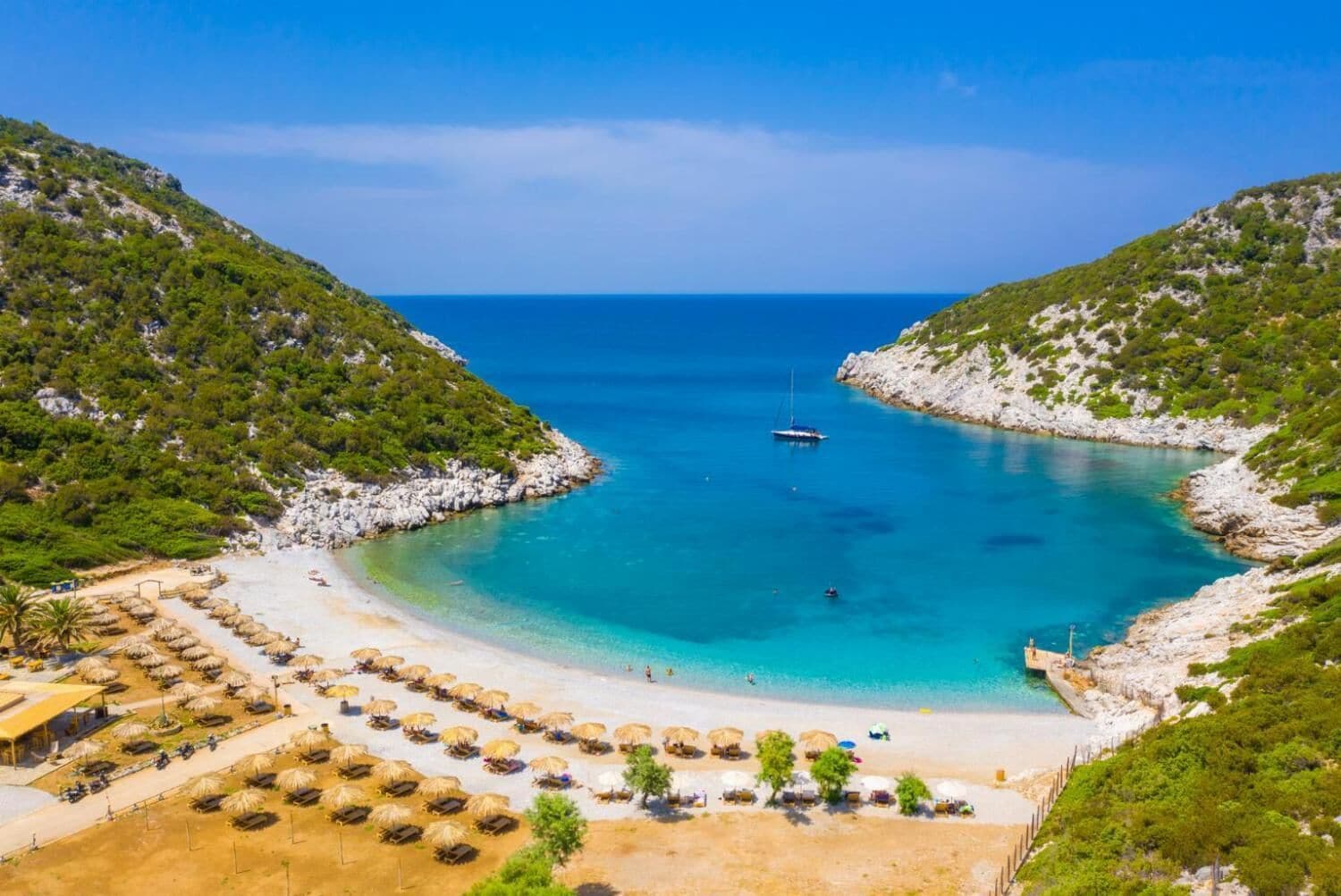 A coastal beach with clear turquoise waters, a boat anchored in the bay, and a row of thatched umbrellas on the sandy shore.