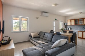 Ein modernes Wohnzimmer mit einer Eckcouch, einem Esstisch mit Stühlen, einer Küchenzeile mit Schränken und Geräten sowie einem an der Wand befestigten Fernseher.