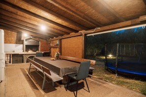 Área de refeições ao ar livre com uma mesa comprida e cadeiras, lareira e um trampolim visível através de uma parede aberta.