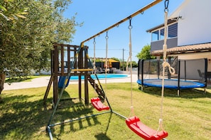 A playground with swings and a slide, a trampoline, and a swimming pool in the background. - Villa Odzakovic - Four Bedroom Villa, Sleeps 8 (Nadin)