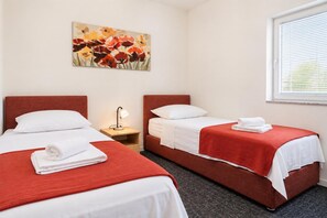 Quarto de hotel com duas camas, mesa de cabeceira com abajur, um quadro na parede e janela com persianas.