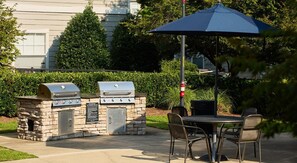Uma estação de churrasco ao ar livre com armários de pedra, uma mesa e cadeiras, com um conjunto de sebe e um edifício ao fundo.