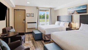 Een hotelkamer met twee bedden, een bureau, een stoel, een televisie en een groot raam met uitzicht op groen.