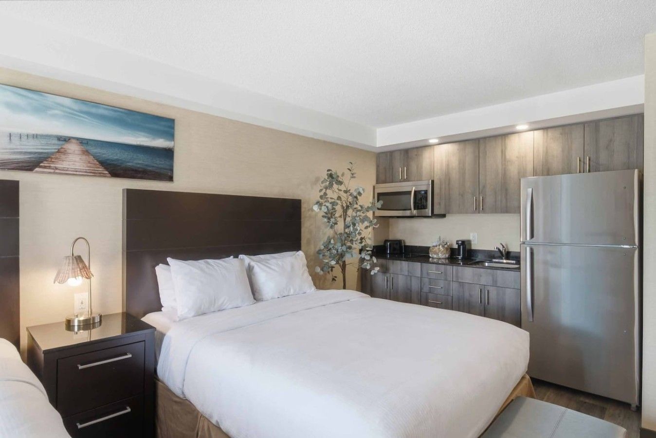 A hotel room with a bed, bedside table, lamp, a painting, a small plant, a kitchenette with a microwave, toaster, coffee maker, and a refrigerator.