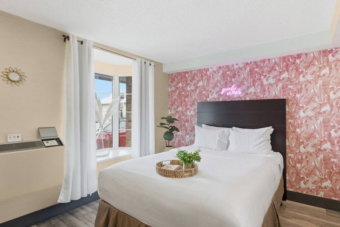 A hotel room with a bed, a small table, a potted plant, and a view of the outside.