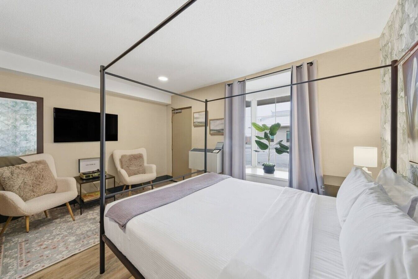 Une chambre d’hôtel avec un lit à baldaquin, deux fauteuils, un bureau avec une lampe et une grande fenêtre avec des rideaux.