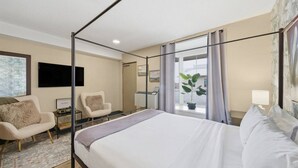 Een hotelkamer met een hemelbed, twee fauteuils, een bureau met lamp en een groot raam met gordijnen.