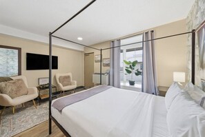Habitación de hotel con cama con dosel, dos sillones, un escritorio con lámpara y un gran ventanal con cortinas.
