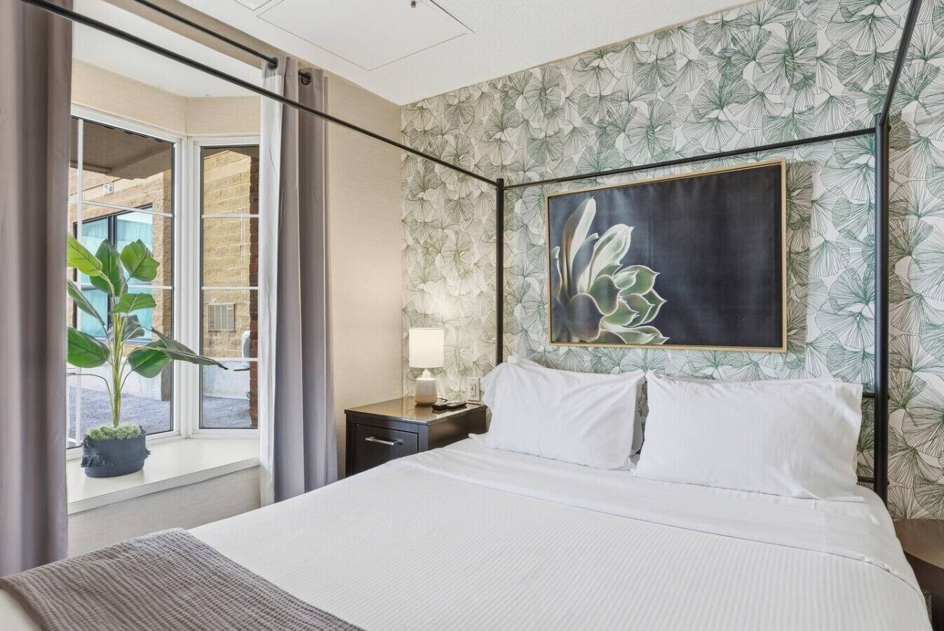 Une chambre d’hôtel avec un lit, une table de chevet, une lampe, une grande fenêtre avec des rideaux et une plante en pot.