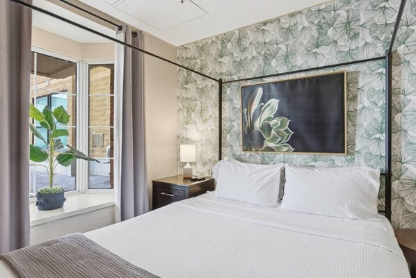 Una habitación de hotel con cama, mesita de noche, lámpara, un amplio ventanal con cortinas y una planta en maceta.