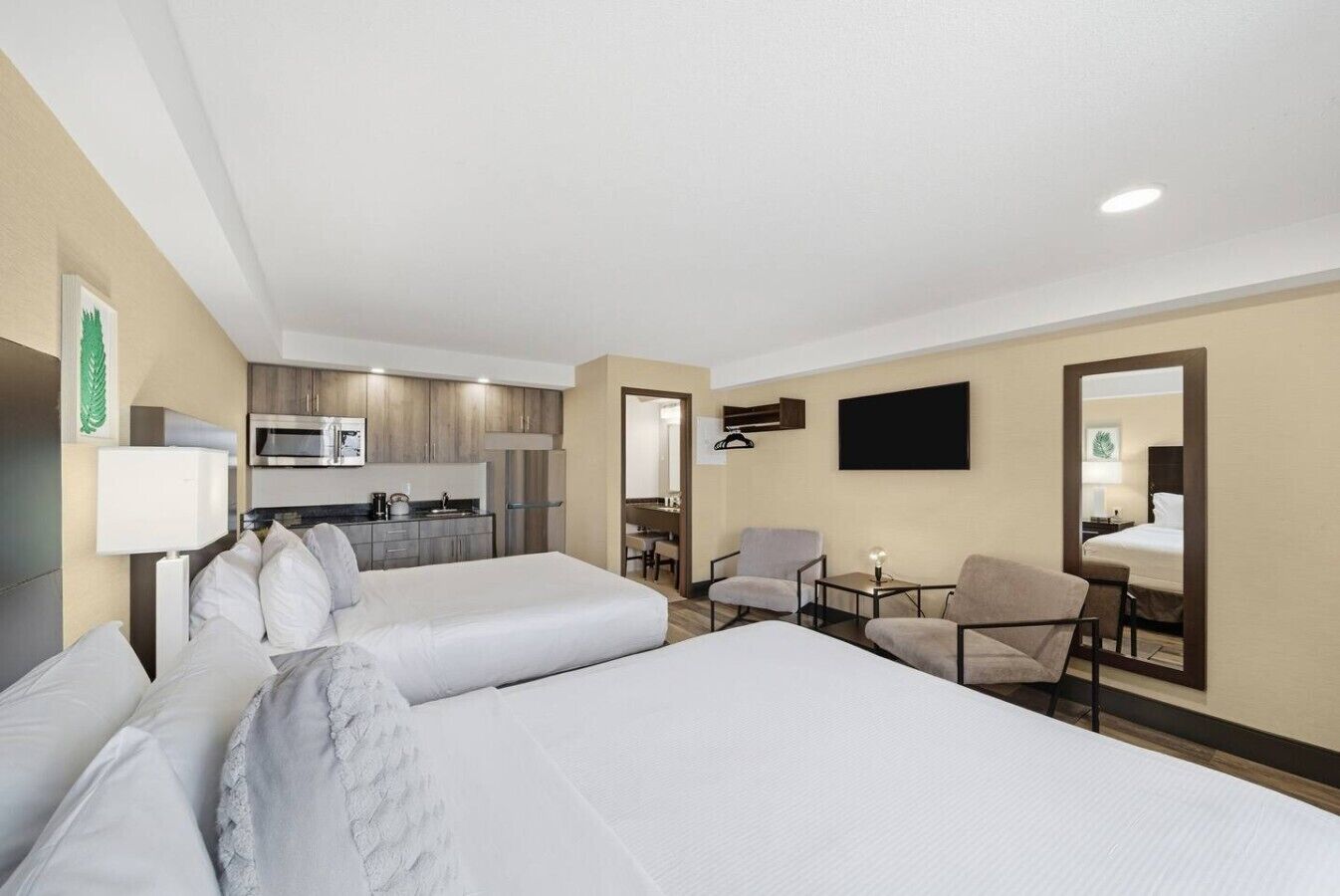 Une chambre d’hôtel avec deux lits, une kitchenette, un coin salon avec des chaises et un téléviseur à écran plat fixé au mur.