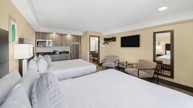 Ein Hotelzimmer mit zwei Betten, einer Küchenzeile, einer Sitzecke mit Stühlen und einem an der Wand befestigten Flachbildfernseher.