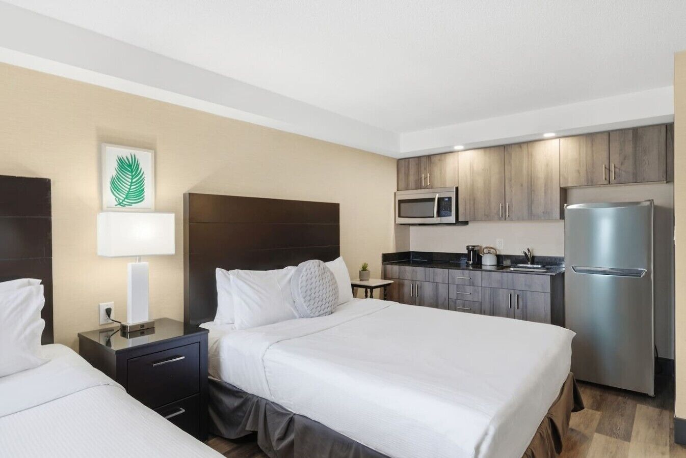 A hotel room with two beds, a kitchenette, a microwave, a refrigerator, and a small table with a lamp.