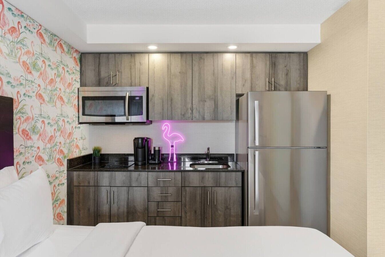 A compact hotel room with a kitchenette, including a microwave, refrigerator, and a neon flamingo sign.
