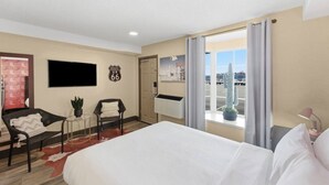 Hotelkamer met een bed, een stoel, een klein tafeltje, een kamerplant, een televisie, een wandklok en een raam met uitzicht op cactussen en een parkeerplaats.