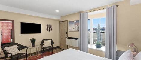 A hotel room with a bed, a chair, a small table, a potted plant, a television, a wall clock, and a window with a view of cacti and a parking lot.