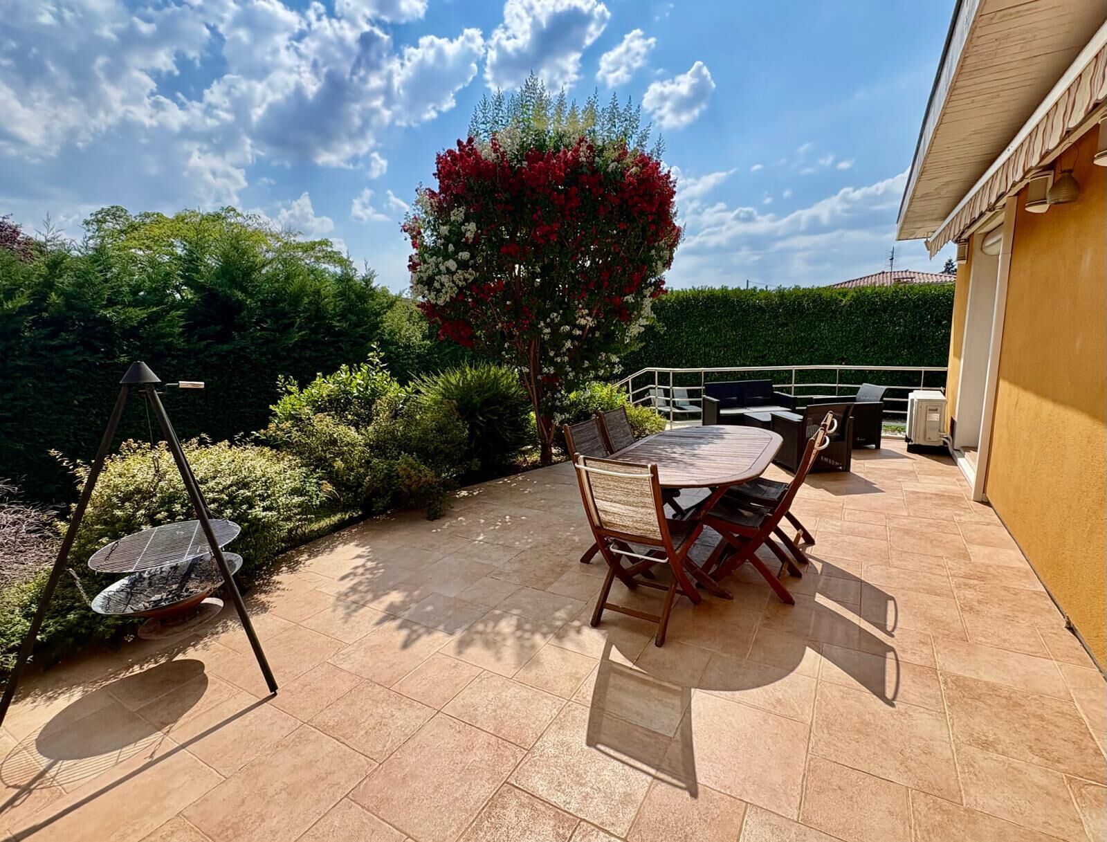Une terrasse extérieure avec une table et des chaises, entourée de verdure et d’une haie.