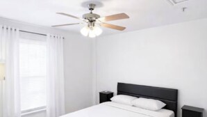 Habitación de hotel con una cama individual, ventilador de techo, mesita de noche y ventana con cortinas.