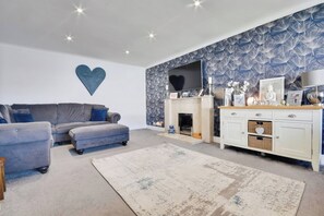 A living room with a grey sofa, a fireplace, a white cabinet, and a patterned wallpaper. - Gralyn Place (Winchelsea)
