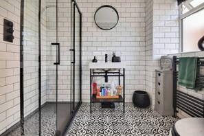 A bathroom with a white tiled wall, a black and white patterned floor, a round mirror, a sink with toiletries, and a green towel on a radiator. - Gralyn Place (Winchelsea)