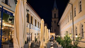 Café ao ar livre com mesas e cadeiras, um edifício com torre do relógio ao fundo e um restaurante com placa.