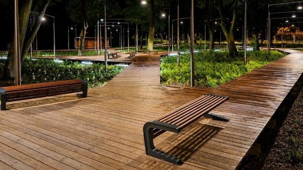 A wooden pathway with benches and street lamps, surrounded by greenery.