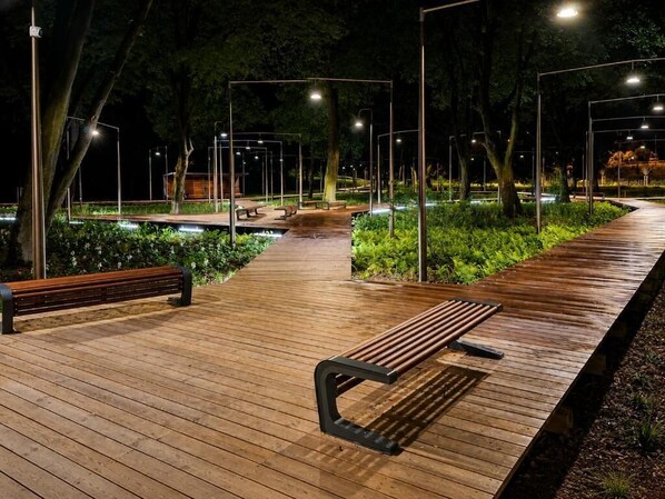 A wooden pathway with benches and street lamps, surrounded by greenery.