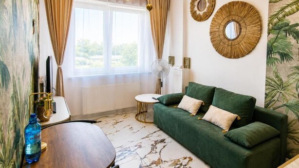 Quarto com sofá verde, espelho redondo, ventilador e janela com cortinas.