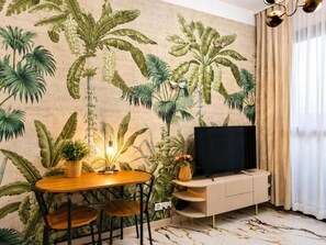 Ein Zimmer mit einer Wandmalerei von tropischen Pflanzen, einem Esstisch mit Stühlen, einem Fernseher auf einem Holzboden und einem Fenster mit Vorhängen.